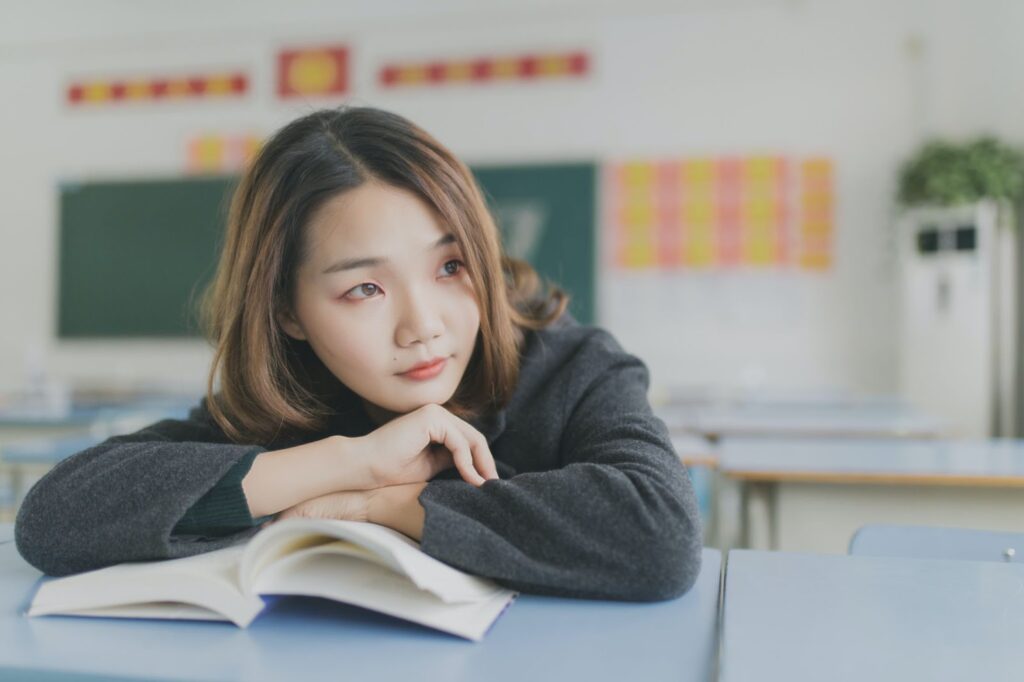 A woman sits with her arms crossed over an open book while in a classroom setting.