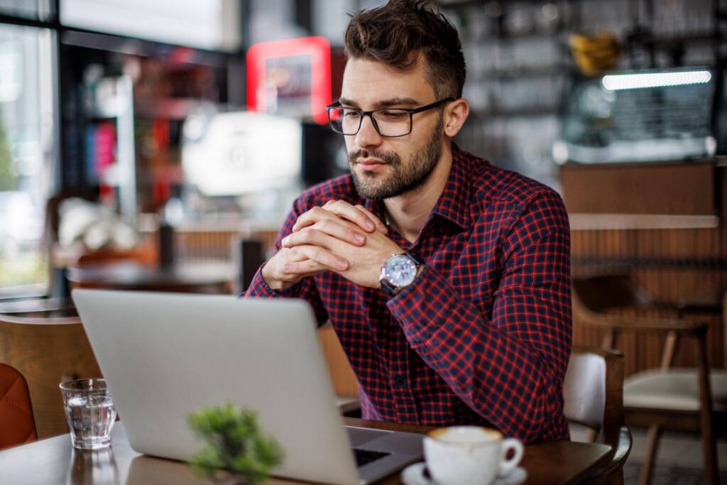 A young man sitting in a coffee shop works at a laptop computer.