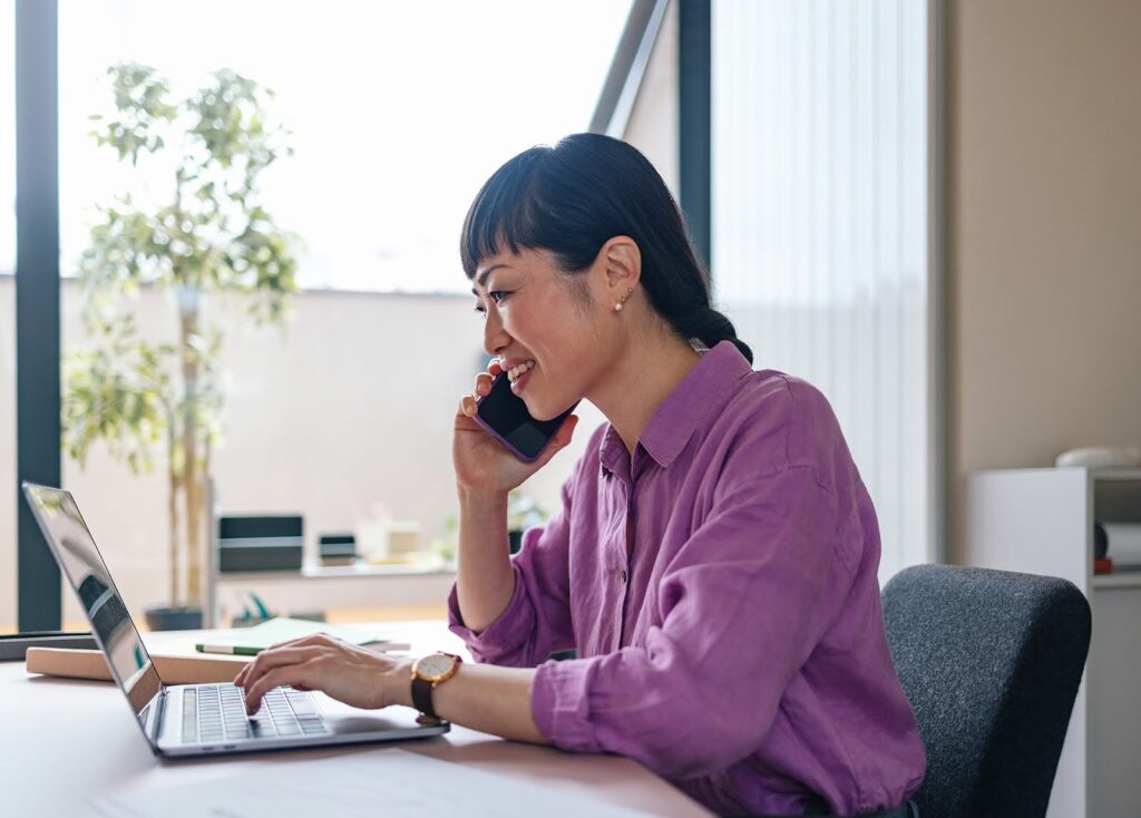 A smiling businesswoman in a bright office setting multitasks, typing at a laptop while engaging in a phone conversation.