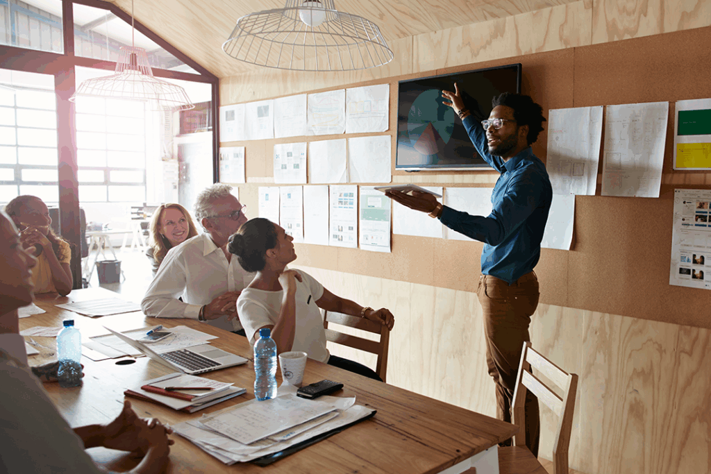 A team convened in a meeting room watches a presenter explain the content of a pie graph projected on a screen behind him.