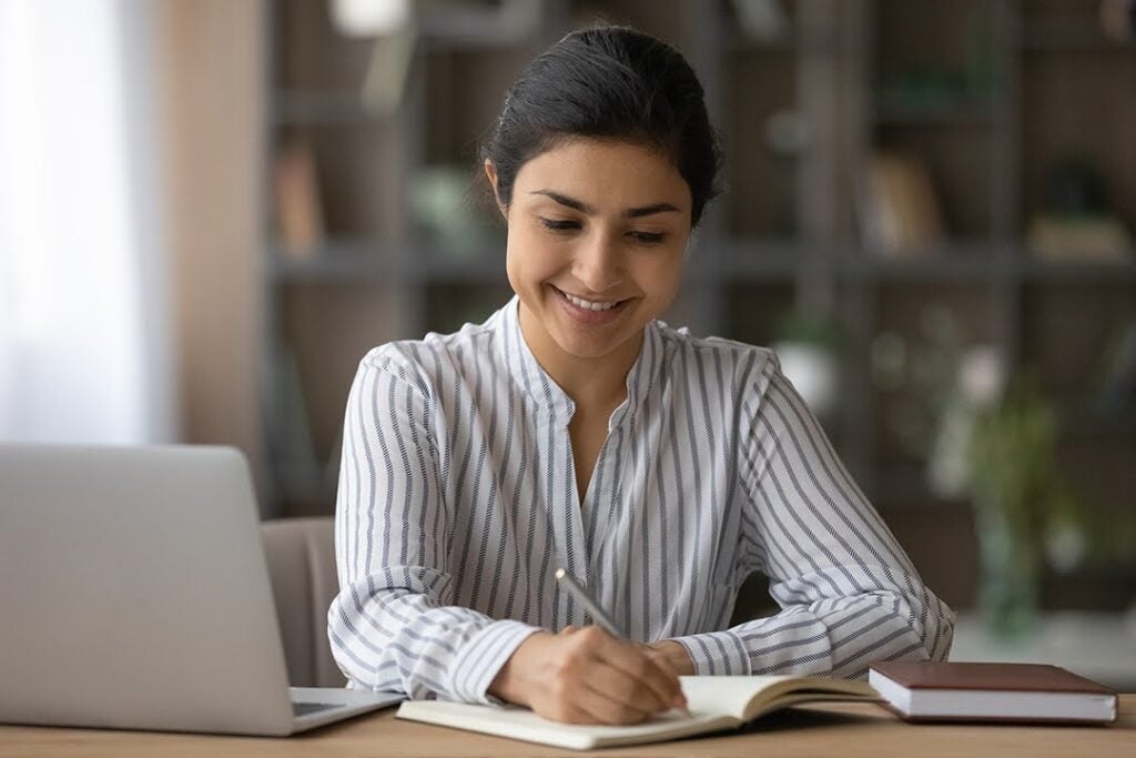 A smiling young woman in a home study setting writes in a notebook; an open laptop sits on the desk before her.