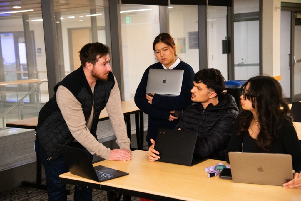 A group of students with laptops converse in a classroom setting.